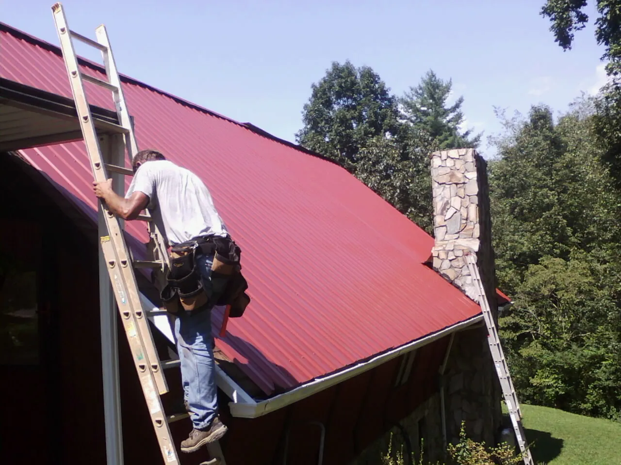 Roofer on a ladder beside a red metal roof and stone chimney — Soffit & Fascia Repair by Roofing Brothers in Arkville